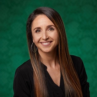 Head shot of Mila wearing a Black deep v button down shirt,  long straight brown highlighted hair, wearing silver hoops, in front of a green background